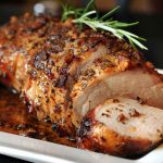 Golden roasted pork loin sliced and served on a platter with fresh herbs and roasted vegetables.