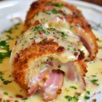 A plate of golden, crispy chicken cordon bleu sliced open to reveal melted Swiss cheese and ham, served with creamy sauce and fresh parsley garnish.