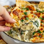A warm dish of baked spinach artichoke dip topped with melted golden cheese, served with toasted bread slices and crackers.