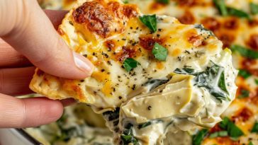 A warm dish of baked spinach artichoke dip topped with melted golden cheese, served with toasted bread slices and crackers.