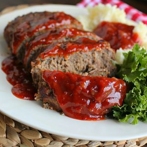 A slice of tender, juicy meatloaf topped with a rich glaze, served with mashed potatoes and green beans on a dinner plate.