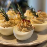 A platter of deviled eggs filled with a creamy yolk mixture, garnished with paprika, chives, and crispy bacon crumbles.