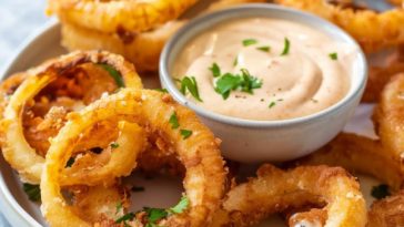 A basket of golden, crispy onion rings served with a creamy dipping sauce in a small bowl, garnished with parsley.