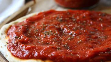 A jar of rich homemade pizza sauce made with tomatoes, garlic, herbs, and olive oil, surrounded by fresh basil and a wooden spoon.