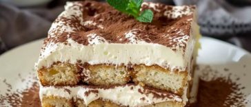 A glass dish of classic Italian tiramisu with layers of espresso-soaked ladyfingers, mascarpone cream, and cocoa powder dusting, garnished with chocolate shavings.