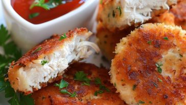 Golden, crispy chicken patties stacked on a plate, served with dipping sauce and garnished with fresh herbs.