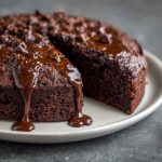 A slice of moist chocolate cake with glossy chocolate frosting, served on a white plate with a fork and a dusting of cocoa powder.