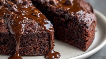 A slice of moist chocolate cake with glossy chocolate frosting, served on a white plate with a fork and a dusting of cocoa powder.