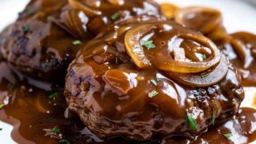 A sizzling hamburger steak smothered in rich brown gravy with caramelized onions, served alongside mashed potatoes and green beans on a rustic dinner plate.