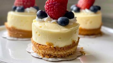 A tray of mini cheesecakes with golden graham cracker crusts, creamy filling, and assorted toppings like berries, chocolate drizzle, and whipped cream.