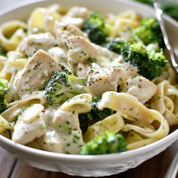 A bowl of creamy Alfredo pasta with tender chicken strips and bright green broccoli florets, topped with Parmesan and fresh herbs.