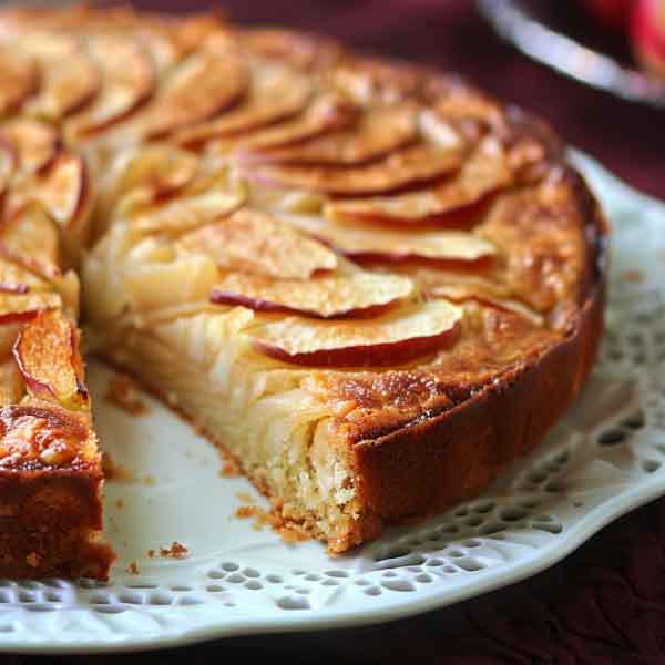 Rustic French apple cake dusted with powdered sugar, served with apple slices and whipped cream on a vintage plate.