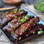 A plate of glistening Korean BBQ ribs garnished with sesame seeds and green onions, served with steamed rice and kimchi on a rustic wooden table.