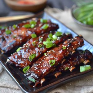 A plate of glistening Korean BBQ ribs garnished with sesame seeds and green onions, served with steamed rice and kimchi on a rustic wooden table.