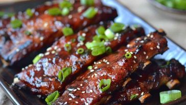 A plate of glistening Korean BBQ ribs garnished with sesame seeds and green onions, served with steamed rice and kimchi on a rustic wooden table.