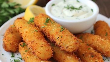 Golden homemade fish sticks served on a plate with lemon wedges, dipping sauce, and a sprinkle of fresh herbs.