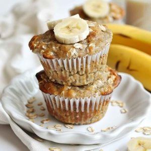 A batch of golden-brown banana oat muffins cooling on a wire rack, with visible oats on top and a sliced banana in the background.