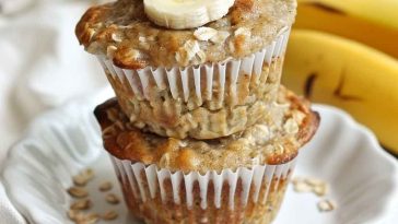 A batch of golden-brown banana oat muffins cooling on a wire rack, with visible oats on top and a sliced banana in the background.