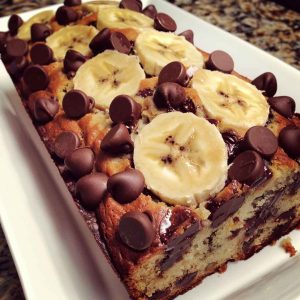 Moist banana chocolate chip bread sliced on a wooden board, with melted chocolate chips and ripe bananas in the background.
