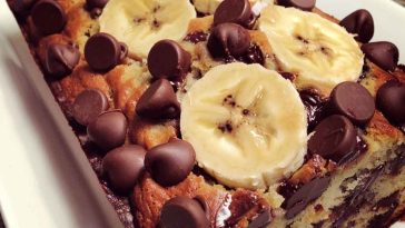 Moist banana chocolate chip bread sliced on a wooden board, with melted chocolate chips and ripe bananas in the background.