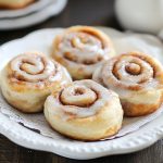 Mini cinnamon rolls glazed with icing, arranged on a plate with cinnamon sticks and powdered sugar.