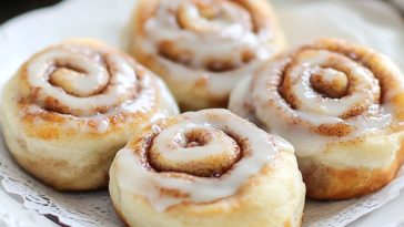 Mini cinnamon rolls glazed with icing, arranged on a plate with cinnamon sticks and powdered sugar.