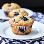 A batch of golden blueberry muffins with juicy berries peeking through, served on a rustic tray with a side of fresh blueberries.