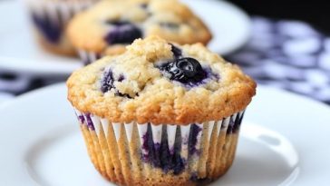 A batch of golden blueberry muffins with juicy berries peeking through, served on a rustic tray with a side of fresh blueberries.