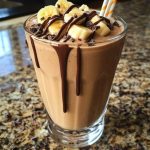 A tall glass of chocolate peanut butter banana shake topped with whipped cream, chocolate drizzle, and sliced bananas.