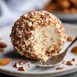 A round cheese ball coated in toasted almonds, served on a platter with crackers, fresh herbs, and grapes.