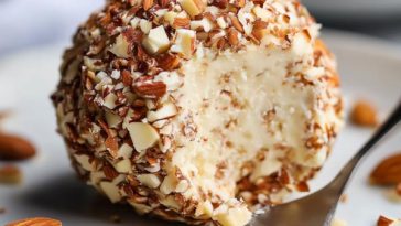 A round cheese ball coated in toasted almonds, served on a platter with crackers, fresh herbs, and grapes.