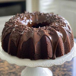 Rich chocolate bundt cake drizzled with glossy ganache, garnished with powdered sugar and berries on a cake stand.