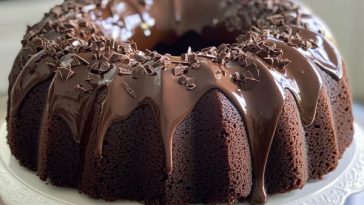 Rich chocolate bundt cake drizzled with glossy ganache, garnished with powdered sugar and berries on a cake stand.