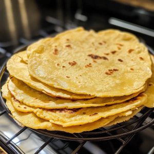 Stack of warm homemade corn tortillas on a rustic plate, soft and golden with a slightly charred surface.