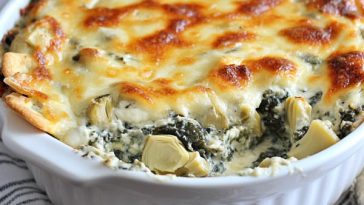 A warm dish of spinach artichoke dip with melted cheese bubbling on top, served with toasted baguette slices and veggie sticks.