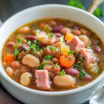 A rustic bowl of ham and bean soup with chunks of ham, white beans, carrots, and herbs in a golden broth, served with crusty bread.