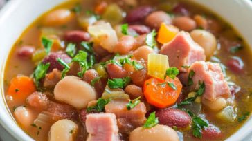 A rustic bowl of ham and bean soup with chunks of ham, white beans, carrots, and herbs in a golden broth, served with crusty bread.