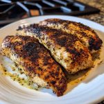 A sizzling blackened chicken breast with charred edges, served over rice and garnished with fresh herbs and lemon wedges.