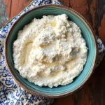 A bowl of freshly made ricotta cheese with a soft, creamy texture, garnished with basil and served with crusty bread.