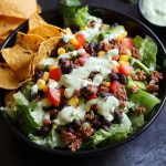 A vibrant taco salad topped with seasoned ground beef, shredded cheese, cherry tomatoes, avocado, and a drizzle of cilantro lime ranch dressing.