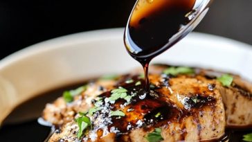 A small glass jar of glossy balsamic glaze with a spoon, surrounded by fresh tomatoes, mozzarella, and basil.