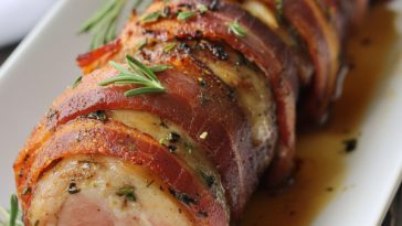 Juicy bacon-wrapped pork tenderloin sliced on a cutting board with a crispy, golden bacon exterior.
