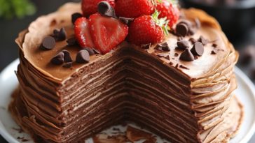 Chocolate mousse crepe cake layered with thin crepes and rich chocolate mousse, topped with cocoa powder and chocolate shavings, served on a cake stand.