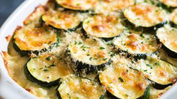 Cheesy zucchini gratin baked golden brown in a casserole dish, layered with sliced zucchini, creamy sauce, and melted cheese, garnished with fresh herbs.