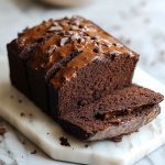 "A loaf of rich brownie bread sliced to reveal its fudgy, chocolatey texture, topped with a dusting of cocoa powder."