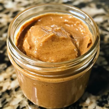 "A jar of creamy homemade peanut butter made with roasted peanuts, displayed with a spoonful beside it."