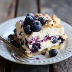 “A layered blueberry cream cheesecake lasagna topped with whipped cream and fresh blueberries in a glass dish.”
