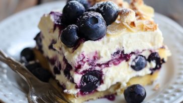 “A layered blueberry cream cheesecake lasagna topped with whipped cream and fresh blueberries in a glass dish.”