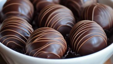 A white bowl filled with round, dark chocolate truffles decorated with fine milk chocolate drizzles, arranged neatly on a wooden surface.