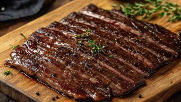 A beautifully cooked and sliced flank steak on a wooden cutting board. The steak appears to be seasoned with cracked black pepper and garnished with fresh thyme, giving it a savory and aromatic finish.